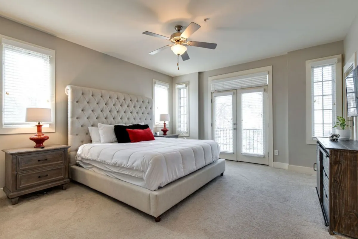 A serene bedroom scene with a plush bed featuring a tufted headboard, a stylish nightstand with a lamp, and sunlight streaming through sliding glass doors. The calm, cozy atmosphere suggests a restful environment ideal for relaxing after a busy day.