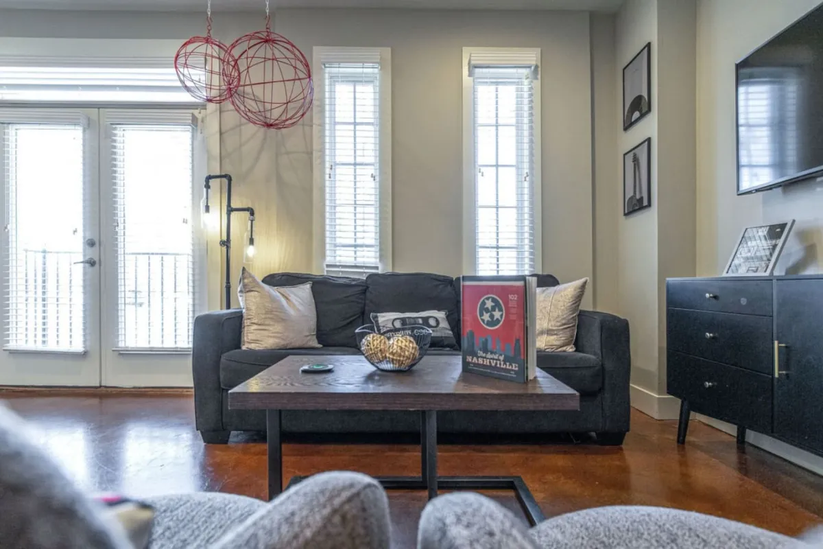The image showcases a bright, inviting living area with a plush sofa, stylish coffee table, and thoughtfully curated decorative accents. Large windows flood the space with natural light, creating an airy and comfortable atmosphere ideal for relaxing, socializing, or savoring a morning coffee. The cozy yet modern decor hints at a perfect blend of comfort and Nashville flair.
