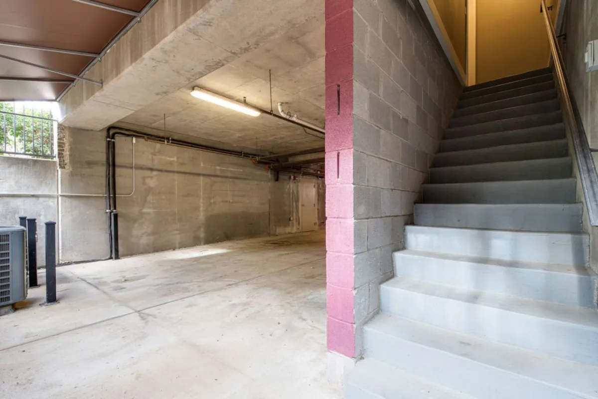 A utilitarian interior garage scene featuring a staircase, exposed concrete walls, and modern lighting. The space is open, practical, and clearly designed for easy access to parking and storage, with a sense of spaciousness and order.