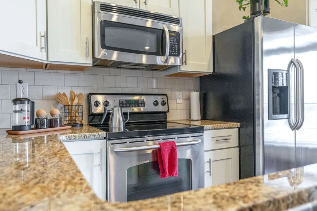The image showcases a modern kitchen with stainless steel appliances, a granite countertop, and a pop of color from a red kitchen towel. Wooden utensils add a homey touch, while the inviting layout suggests a convenient, well-equipped space for preparing meals or entertaining guests.