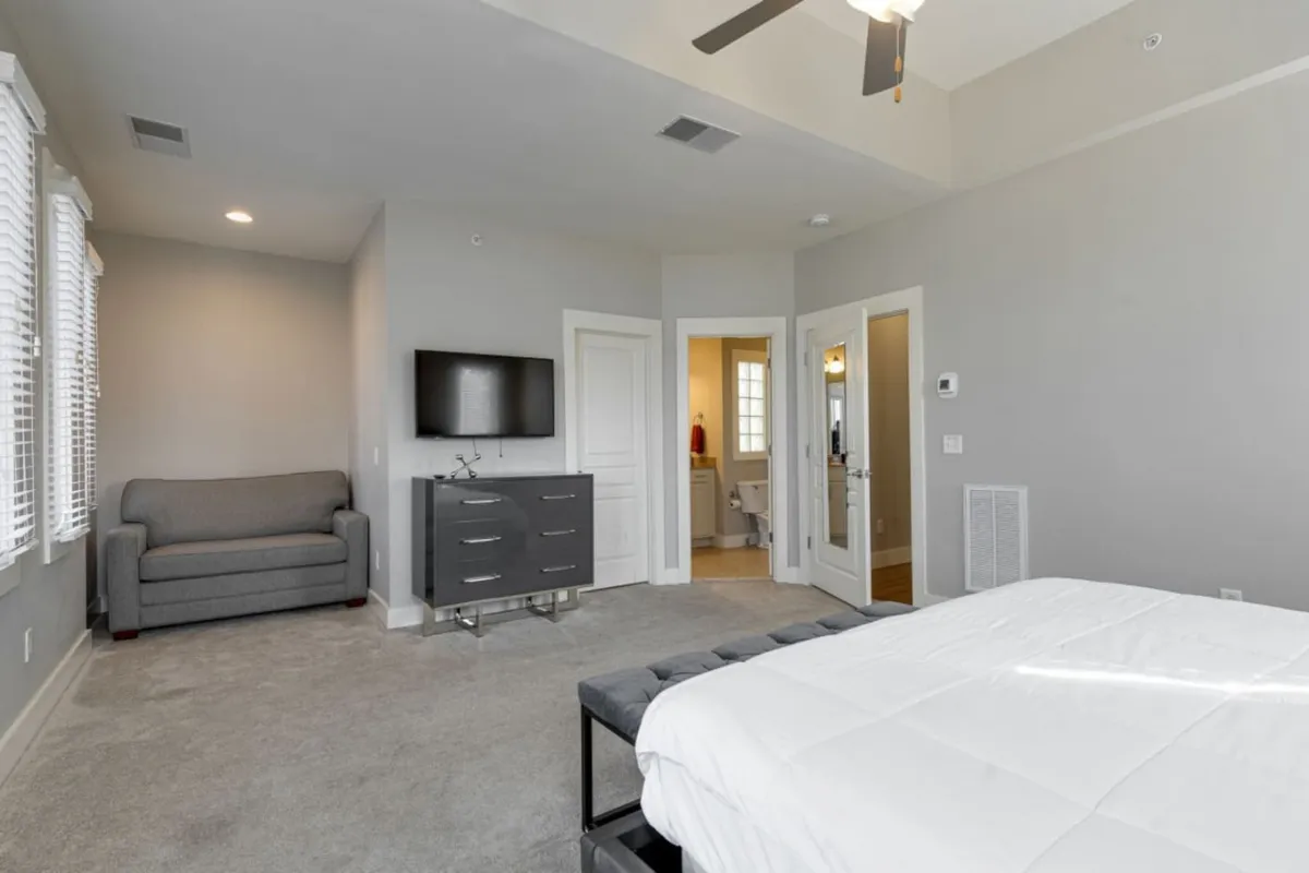 A spacious bedroom featuring a king bed, a large sleeper sofa, a dresser, a television, and a bathroom door visible in the background. The room is staged for both relaxation and functionality, suitable for families or groups needing flexible sleeping arrangements.