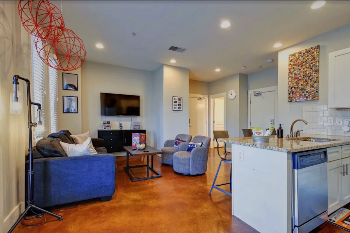 This image showcases a stylish living room with a plush sofa, coffee table, modern artwork, and a visible kitchen area in the background. The space feels cozy and contemporary, featuring an entertainment center and inviting decor, ideal for relaxing or socializing.