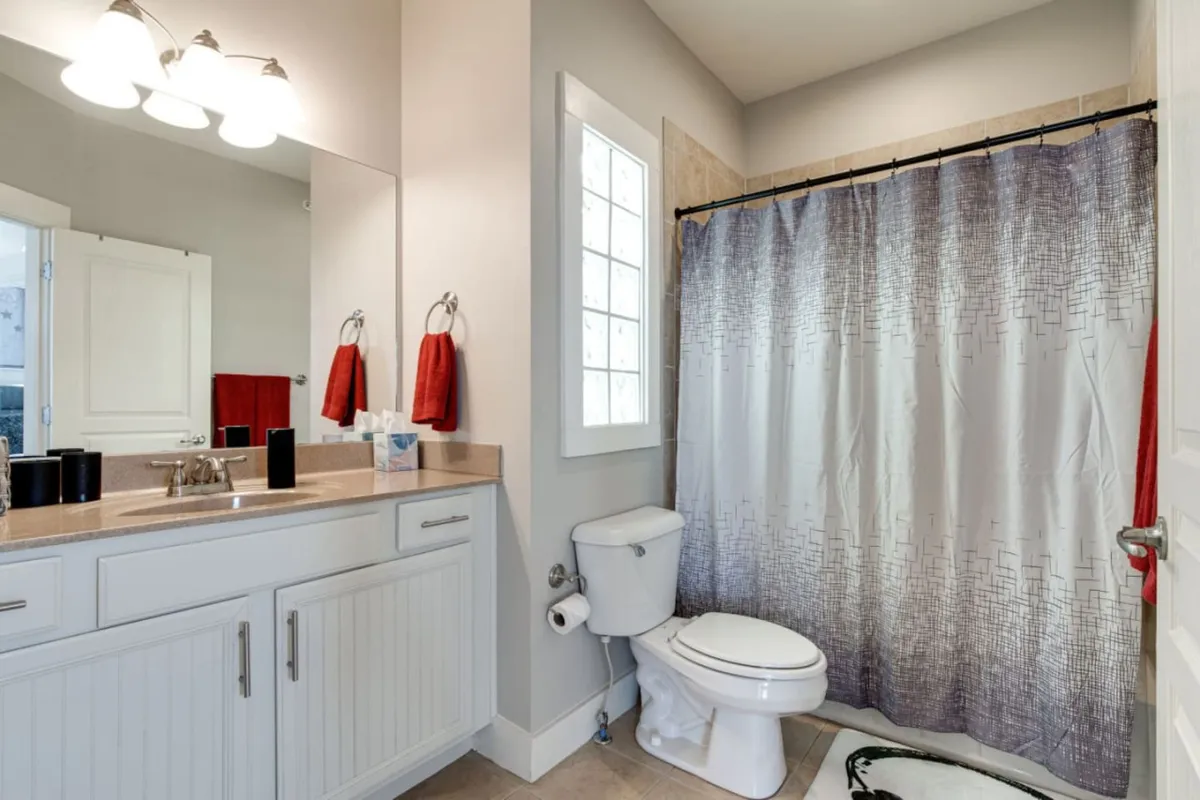 A fresh, tidy bathroom is depicted with a modern vanity, shower curtain, and neatly arranged red towels on a rack. The scene suggests a private, peaceful space for relaxation and self-care after a long day at work or exploring the city.