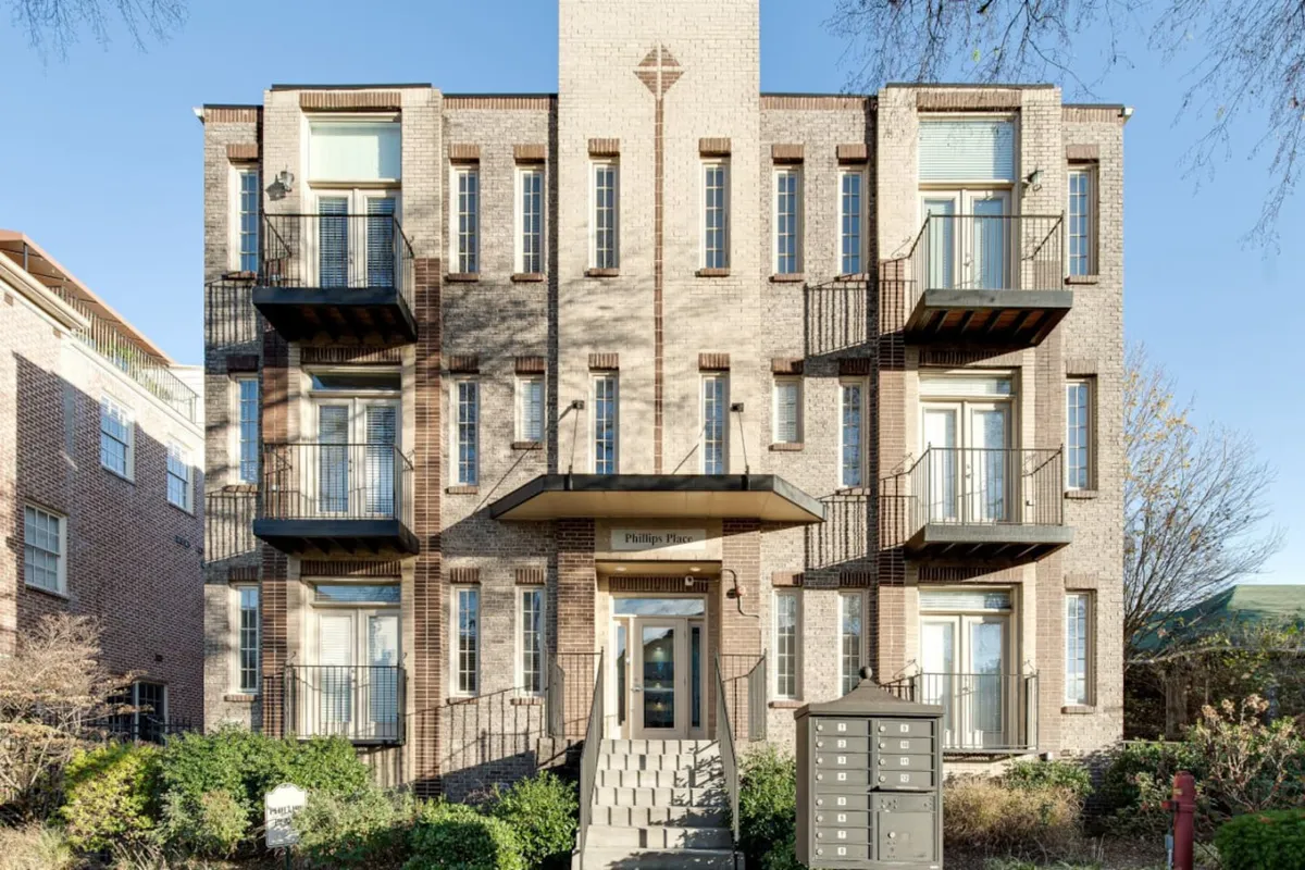 This image features the building's inviting exterior facade, with balconies overlooking a quiet neighborhood street. The entrance door and mailboxes are visible, as are people enjoying time together on the balconies and walking in the neighborhood, creating a lively yet welcoming scene.