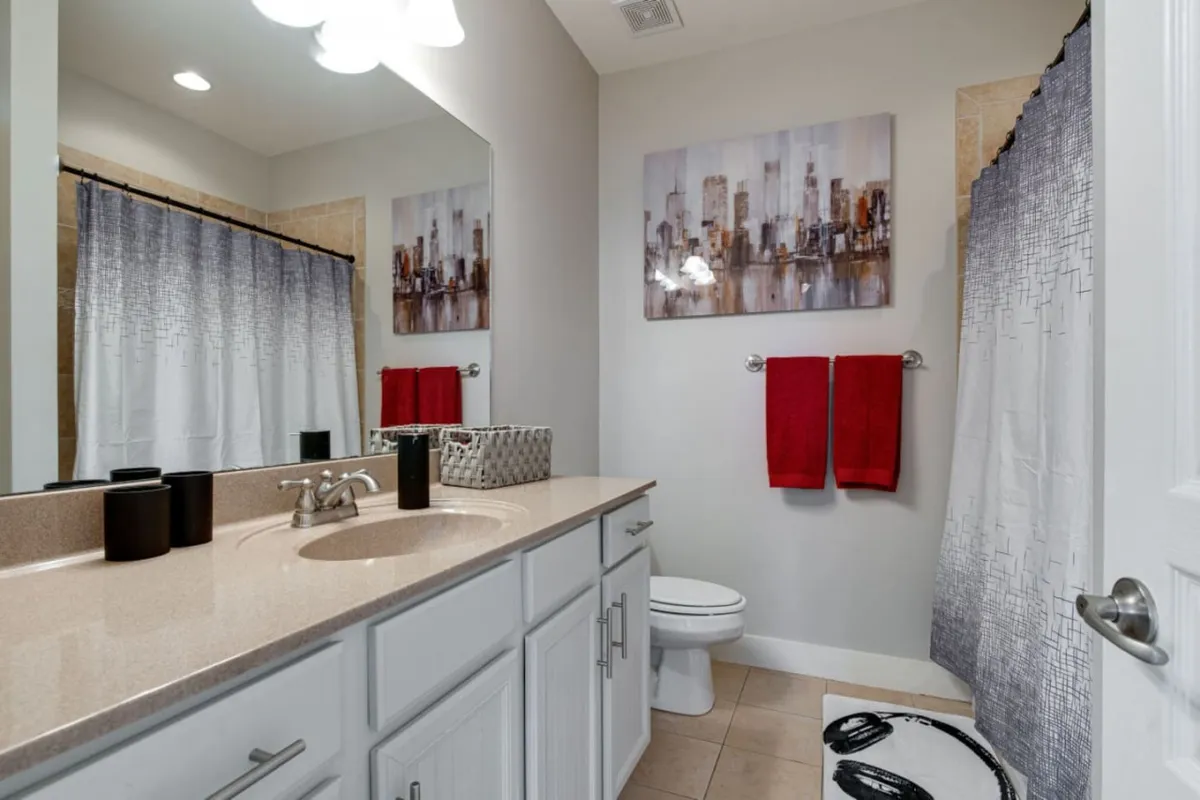 A bright, modern bathroom scene with chic artwork, vibrant red towels, a spacious countertop, and stylish light fixtures. The clean and inviting design emphasizes comfort and luxury in the ensuite bath space.